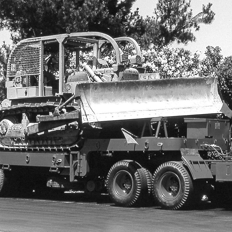 A black and white photo of a large bulldozer on the back of a low bed equipment trailer ready for deployment to a fire.